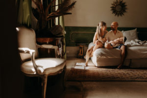 Photo de famille dans une chalbre en provence. Ils tiennent leur bébé dans les bras. Ils sont habillés de façon estivale, dans des couleurs et matières intemporelles