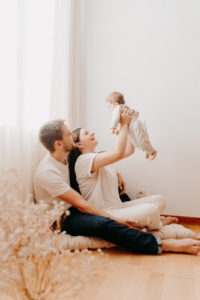 Un couple de jeune parent, chez eux, habillés de couleurs claires près d'une fenêtre