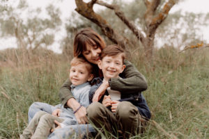 Une maman et ses deux fils assis dans l'herbe. Leurs tenues est en accord avec la nature et le confort. Ils rigolent