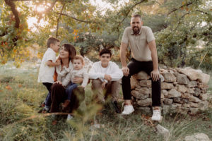Photo de famille, ils sont assis sur un muret de pierres sèches, habillés de façon décontractée dans des tons similaires et des chaussures adaptées pour une séance photo de famille en provence